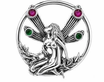 May include: A circular silver-toned brooch featuring a central figure with wings, accented by two round red stones at the top and two round green stones on the sides. The design is enclosed within a polished metal ring, showcasing intricate detailing.