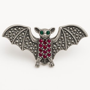 May include: A silver-coloured bat brooch with outstretched wings. The wings are detailed with small, dark grey stones. The bat's body is adorned with red gemstones, and it has green eyes. A unique and detailed accessory.