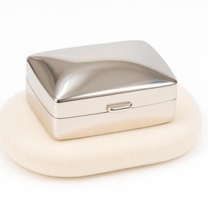 May include: A rectangular silver-toned metal box with a smooth, polished finish and a slightly domed lid. The container features a small, centered latch on the front edge, providing a clean and reflective surface appearance for this storage item.