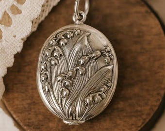 Può includere: Ciondolo ovale in argento con un rilievo dettagliato di fiori e foglie di mughetto. Il ciondolo ha un anello nella parte superiore per una catena. Lo sfondo è una superficie di legno.