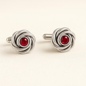 May include: A pair of silver cufflinks with a rose design. Each cufflink features a swirling, textured silver pattern surrounding a round, deep red gemstone. The cufflinks are displayed on a neutral background.