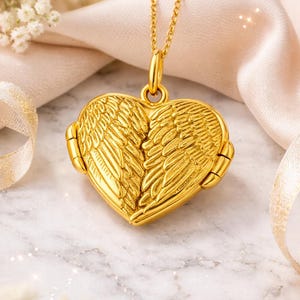 Peut inclure: Un collier avec un pendentif en forme de cœur doré, orné d'ailes d'ange. Le pendentif est suspendu à une chaîne délicate. Le médaillon présente des détails en forme de plumes et une fermeture à fermoir. Le fond comprend des fleurs blanches et une surface en marbre.