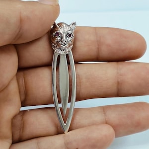 May include: A silver-colored metal bookmark featuring a detailed cat's head with red gemstone eyes. The bookmark has a sleek, elongated design, perfect for marking pages in books. The cat's face is intricately sculpted, adding a touch of elegance.