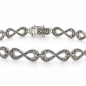 Op de afbeelding: Een zilverkleurige armband met een herhalend patroon van oneindigheidssymbolen. Elke oneindigheidsschakel is versierd met kleine, donkere, gefacetteerde steentjes. De armband heeft een rechthoekige baksluiting die ook met steentjes is bezet.