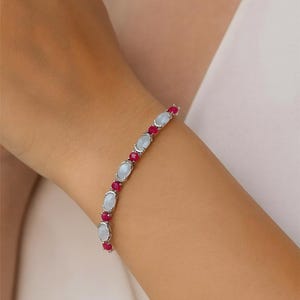 May include: A silver bracelet featuring alternating oval-shaped, light blue gemstones and small, round, red gemstones. The bracelet is displayed on a wrist, showcasing the jewelry's design and color contrast.