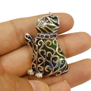 Puede incluir: Un broche de plata en forma de gato con esmalte verde y azul y una fila de piedras preciosas púrpuras alrededor del cuello.