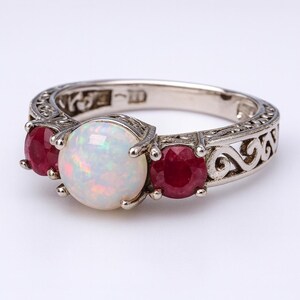 Puede incluir: Anillo de plata con una ópalo iridiscente grande y dos rubíes rojos. El anillo tiene un diseño de filigrana ornamental a lo largo de la banda. Las piedras preciosas están engastadas en una montura de garras. El diseño del anillo sugiere un estilo vintage o antiguo.