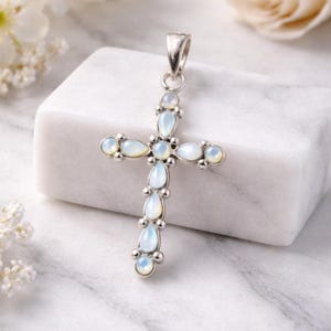 Puede incluir: Un colgante de plata con forma de cruz, adornado con piedras opalescentes en forma de lágrima. La cruz tiene un lazo para una cadena y está sobre una superficie de mármol blanco. Joyería religiosa.
