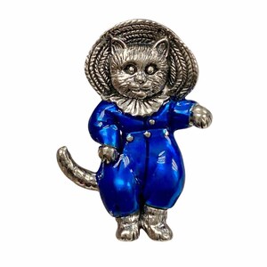 Könnte beinhalten: Silberfarbene Katzenbrosche mit blauem Emaille-Outfit und gewebtem Hut. Die Katze steht mit einer ausgestreckten Pfote. Die Brosche hat ein detailliertes Design und eine Vintage-Ästhetik.