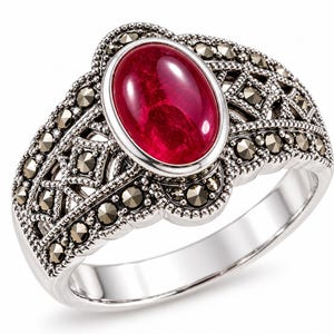 May include: A silver ring featuring a large, oval-shaped, deep red cabochon stone at the centre. The band and setting are decorated with intricate metalwork and numerous small, faceted, dark grey marcasite stones arranged in a geometric pattern.