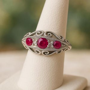 May include: A silver ring with an ornate filigree design. The ring features three vibrant red gemstones, with two smaller stones flanking a larger central stone. The ring is displayed on a white ring holder.