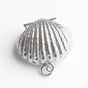 Może przedstawiać: Srebrny wisiorek w kształcie muszli przegrzebka. Wisiorek ma teksturowaną powierzchnię z pionowymi żebrami i małą pętlą na dole. Wisiorek jest prawdopodobnie wykonany z metalu.