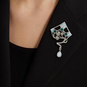 Puede incluir: Broche plateado con diseño de cometa, con detalles de esmalte turquesa y blanco y tres gemas turquesas. Un colgante en forma de lágrima cuelga debajo. El broche está prendido a una blazer negra.