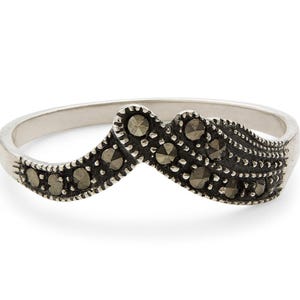 Könnte beinhalten: Ein silberner Ring mit einem wellenförmigen Design. Der Ring hat kleine, dunkle Steine, die in das Silberband eingesetzt sind. Die Steine sind in einem dekorativen Muster angeordnet und verleihen dem Schmuck Eleganz.