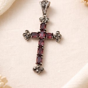 Puede incluir: Colgante de cruz plateada con piedras preciosas ovaladas de color rojo intenso. La cruz presenta detalles ornamentales en la fianza y en los extremos de los brazos. El colgante se muestra sobre una tela color crema.