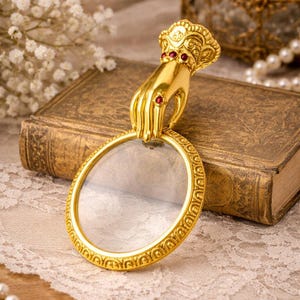 Pode incluir: Uma lupa dourada com uma alça decorativa em forma de mão. A mão é adornada com joias vermelhas. A lupa tem uma moldura ornamentada e padronizada e uma lente de vidro transparente. A estética geral é vintage e elegante.