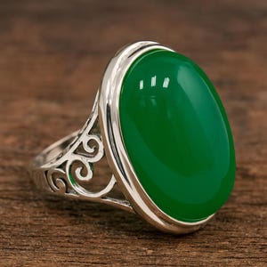 May include: A silver ring with a large, oval, glossy green gemstone. The ring has a decorative filigree design on the sides and a polished silver band. The gemstone is set in a smooth bezel. The ring is on a wooden surface.