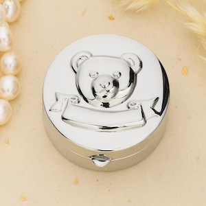 Pode incluir: Uma caixa de recordações redonda em metal prateado com um design de urso de peluche em relevo na tampa. O urso está posicionado acima de uma área de faixa lisa e vazia, adequada para gravação. A caixa tem um acabamento polido e reflexivo.