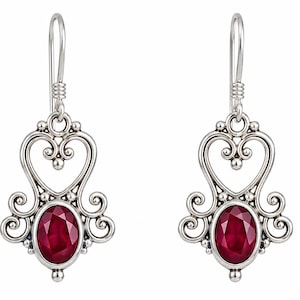 Puede incluir: Un par de pendientes colgantes de plata con una piedra preciosa de color rojo oscuro de corte ovalado engastada en un marco de metal pulido. El diseño incluye intrincados adornos de volutas y filigrana sobre la piedra.