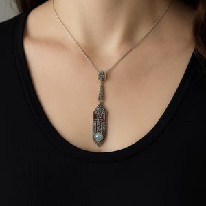 Puede incluir: Un collar con colgante plateado con un diseño geométrico. El colgante presenta una piedra central azul claro, detalles en rojo y pequeños adornos brillantes. El collar se lleva sobre un fondo negro.