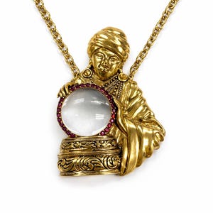 Peut inclure: Collier pendentif doré représentant une figure tenant une boule de cristal. La figure porte un turban et une robe, la boule de cristal étant encadrée de pierres précieuses rouges. Le pendentif est suspendu à une chaîne dorée.
