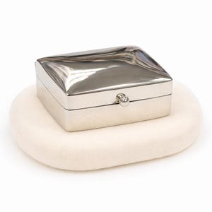 May include: A rectangular silver-toned metal box with a polished, reflective lid and a small spherical knob closure on the front. The box features clean, straight sides and a slightly domed top surface that catches light, creating a smooth finish.