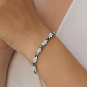 May include: A silver bracelet featuring oval-shaped opals and emeralds. The gemstones are set in a row, alternating between the opals and emeralds. The bracelet is worn on a wrist.