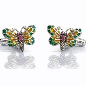 May include: A pair of butterfly cufflinks. The cufflinks feature a silver-toned metal base with a butterfly design. The butterfly wings are green, yellow, and orange, with a small, oval, pink gemstone in the center. The cufflinks are displayed on a white surface.