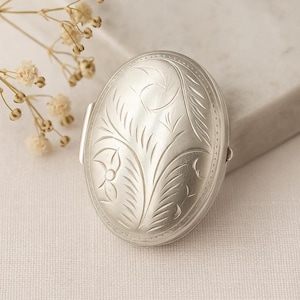 Può includere: Medaglione ovale in argento con un design floreale inciso. Il medaglione ha una chiusura a cerniera e un motivo dettagliato di foglie e fiori. Il metallo ha una finitura lucida e il design è centrato sulla parte anteriore.
