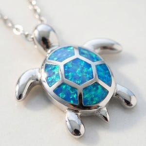 Op de afbeelding: Zilverkleurige schildpendantketting met een levendig blauw en groen opaalachtig middenstuk. De schildpadschild is gesegmenteerd en de ketting is delicaat.