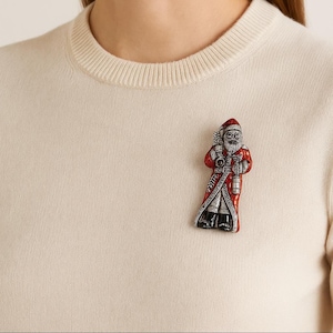 May include: A Santa Claus brooch with a red coat, black boots, and a white beard. The brooch has silver and black accents and is attached to a cream-coloured jumper. The Santa Claus figure is holding a lantern and a bag.