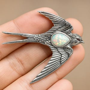 May include: A detailed silver-toned bird pendant with outstretched wings and a teardrop-shaped opal stone. The bird's feathers are intricately carved, and it has a small red gemstone eye. The pendant is held in a hand.