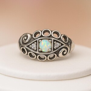 May include: A silver ring with an opal center stone and two smaller accent stones. The ring features an ornate design with swirls and circular details. The opal has a square shape and a light, iridescent color.