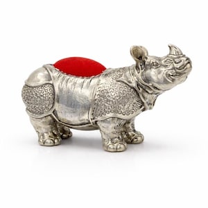 May include: A silver-coloured rhino-shaped pin cushion with a red cushion top. The rhino has detailed textured skin and is standing on four legs. The pin cushion is a decorative sewing accessory.