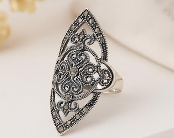 Puede incluir: Anillo plateado con un diseño alargado y ornamentado. El anillo presenta un motivo floral central y está adornado con pequeñas piedras oscuras. El intrincado trabajo de filigrana y la forma general le dan un aspecto vintage o antiguo.