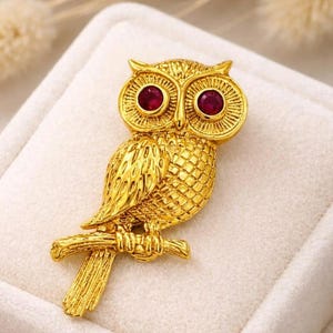May include: A gold-coloured owl brooch with red gemstone eyes. The owl is perched on a branch, with detailed feather and texture patterns. The brooch is set against a white background.