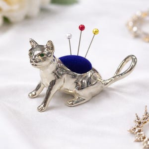 Op de afbeelding: Een zilveren katvormig speldenkussen met een blauw fluwelen kussen en kleurrijke spelden. De kat heeft groene ogen en een gekrulde staart. De spelden zijn rood, geel en wit. Het item staat op een wit oppervlak.