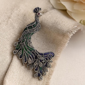 May include: A silver-toned peacock brooch with green and blue enamel detailing. The brooch features a detailed design with marcasite accents and a red gemstone eye. The peacock is posed on a neutral-colored fabric.