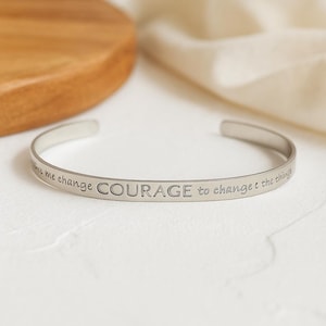 May include: A silver-colored cuff bracelet with the engraved words "COURAGE to change the things". The bracelet is open-ended and sits on a white surface. A wooden circular object is in the background.