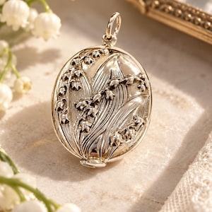 May include: An oval silver locket featuring a detailed embossed design of lily of the valley flowers and leaves on the front surface. The locket has a small circular bail at the top for attachment and a polished metallic finish with dark accents.