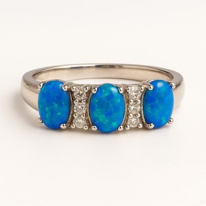 May include: A silver ring with three oval, bright blue opal gemstones. Small, clear accent stones are set between the opals. The ring has a polished finish, and is set against a white background.