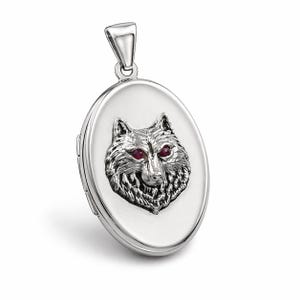 May include: Silver oval locket pendant with a wolf head design and red gemstone eyes. The pendant has a bail for a chain and a hinged design. A decorative piece of jewellery.