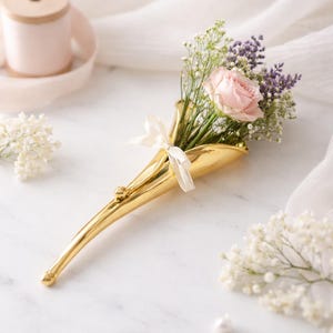 Puede incluir: Un jarrón de metal dorado contiene un pequeño ramo de flores, que incluye una rosa rosa, ramitas moradas y paniculata blanca. Una cinta blanca está atada alrededor del jarrón. El jarrón está sobre una superficie de mármol blanco.