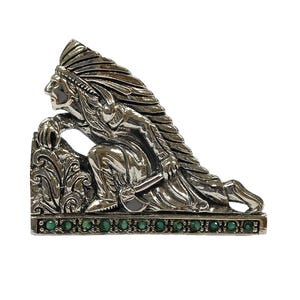 Puede incluir: Escultura de metal plateado que representa una figura nativa americana arrodillada, con un tocado de plumas. La base está adornada con pequeñas piedras preciosas verdes redondas. La figura es detallada, sobre fondo blanco.