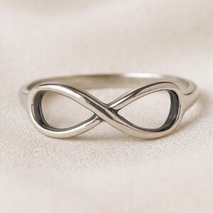May include: A silver ring featuring an infinity symbol design. The ring, made of a smooth, shiny metal, is displayed on a white, textured surface. The infinity symbol is a continuous loop, representing eternity.