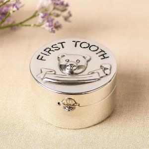Könnte beinhalten: Silberfarbene, runde Andenkenbox mit der Aufschrift "FIRST TOOTH" auf dem Deckel. Der Deckel zeigt ein Teddybären-Design und ein Banner. Die Box hat einen Klappverschluss, wahrscheinlich für den ersten Zahn eines Babys.