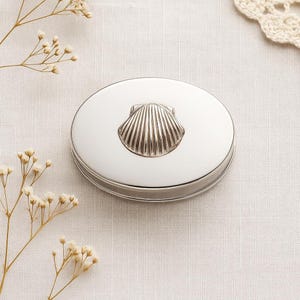 May include: A silver-colored, oval-shaped trinket box with a seashell design on the lid. The box is made of a shiny metal and sits on a white surface. The seashell is detailed with vertical lines.