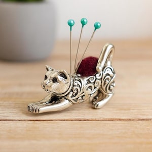 May include: A silver-colored cat-shaped pin cushion with a burgundy cushion and three teal-headed pins. The cat is in a stretching pose, with intricate scrollwork detailing its body. A decorative sewing accessory.