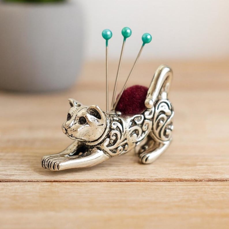 May include: A silver-colored cat-shaped pin cushion with a burgundy cushion and three teal-headed pins. The cat is in a stretching pose, with intricate scrollwork detailing its body. A decorative sewing accessory.