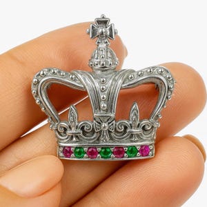 Könnte beinhalten: Silberfarbene Kronenbrosche mit detailliertem Design. Die Brosche ist mit roten und grünen Edelsteinen besetzt. Die Krone hat ein Kreuz an der Spitze und dekorative Verzierungen.